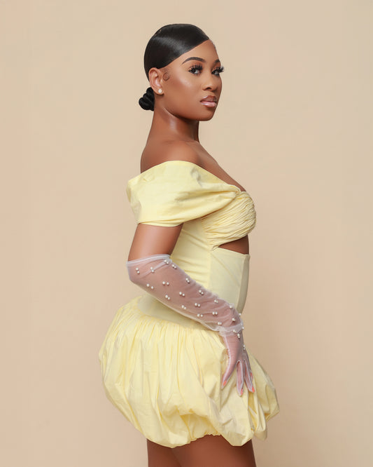 Woman wearing a yellow off-shoulder dress with sheer sleeves against a beige background