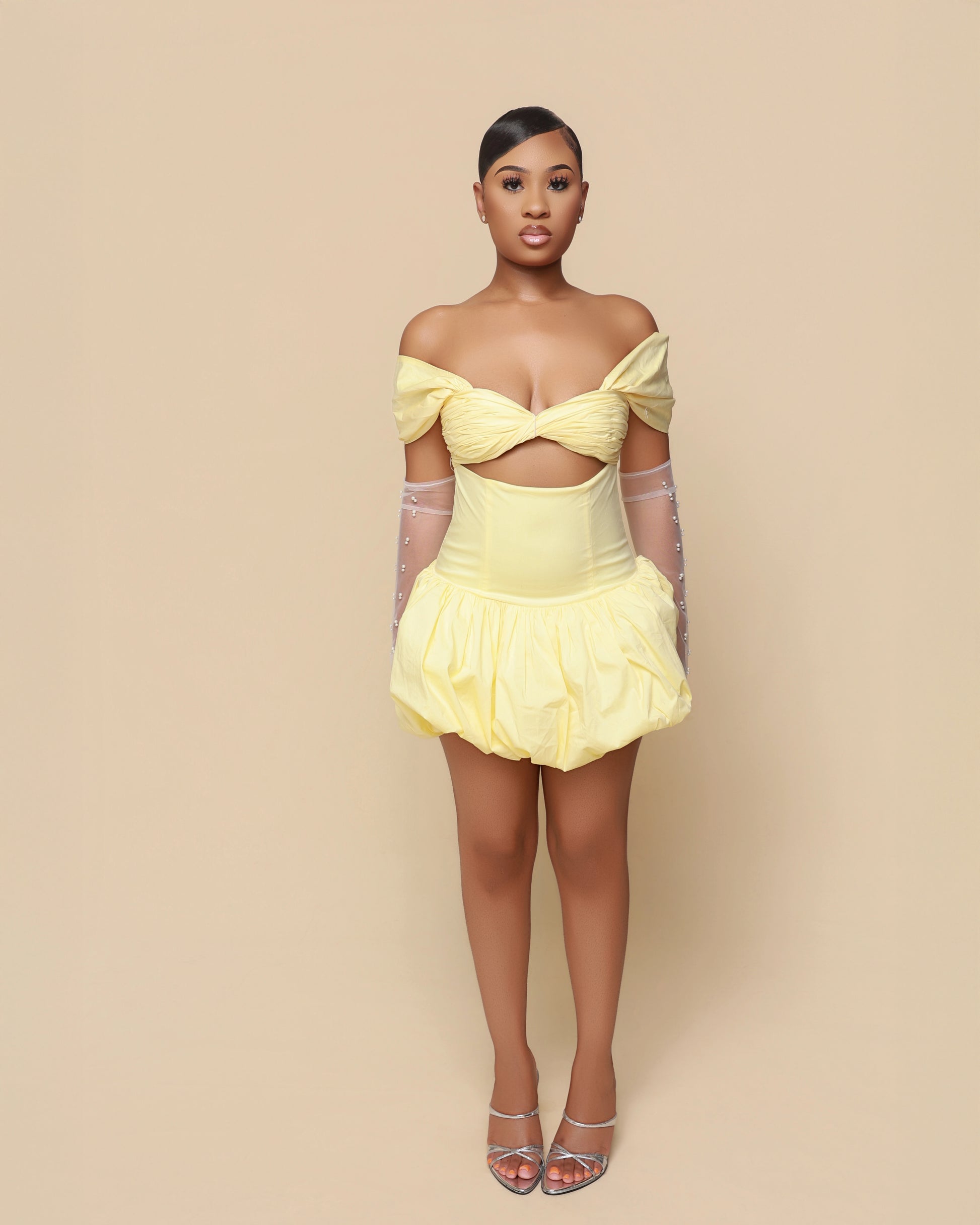 Woman wearing a yellow off-shoulder dress with sheer sleeves against a beige background